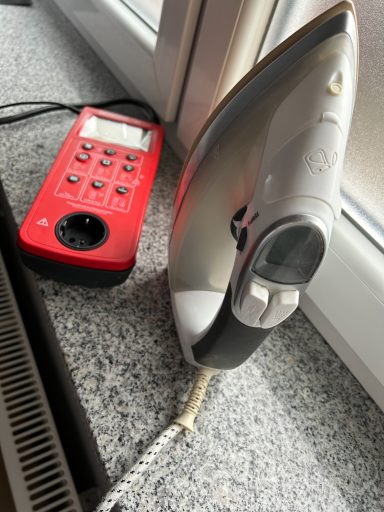 Ein Bügeleisen und eine rote Steckdosenleiste auf einem Tisch neben einem Fenster.