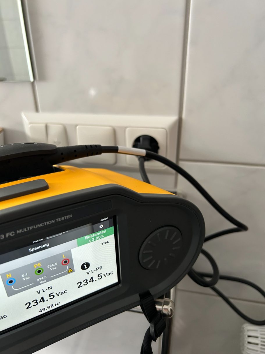 E-Check einer Wohnung Ein gelbes Gerät mit Display und Stecker, das an eine Wandsteckdose angeschlossen ist.