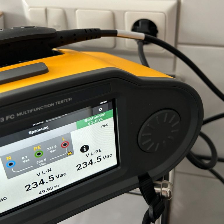 Ein gelbes Messgerät zeigt Spannung und Stromstärke an, angeschlossen an eine Steckdose.